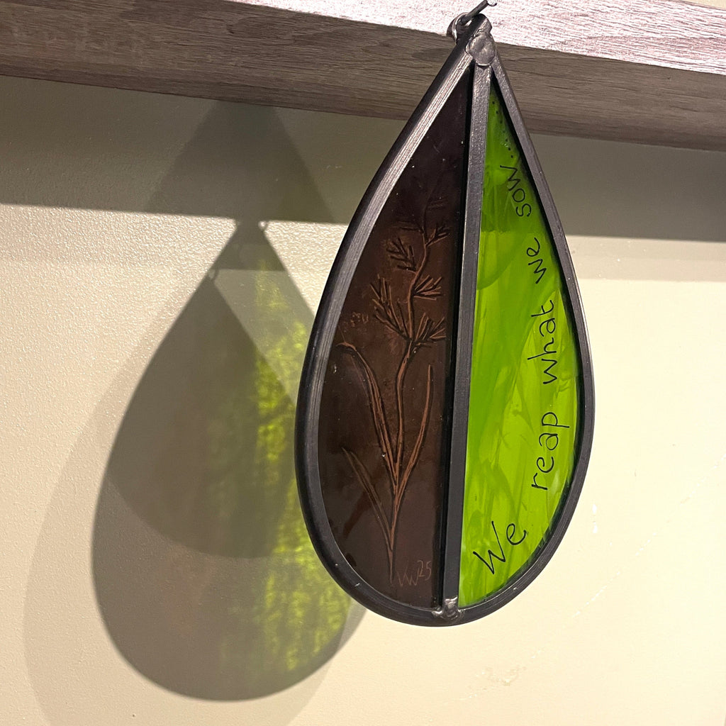 handmade stained glass tree spinner / sun catcher, green and purple glass with hand painting of flower and the words, 'we reap what we sow' on the glass, traditional stained glass painting, with lead, hanging against white wall