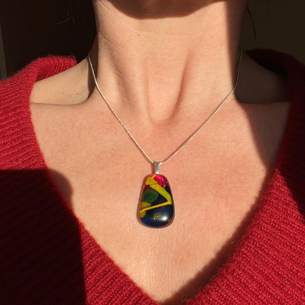 Person wearing a colorful pendant necklace with a red sweater, hand-crafted fused glass pendant. colourful fused glass, rich cherry red, yellow, mustard, green. on sterling silver snake chain, complete one-off, only one available