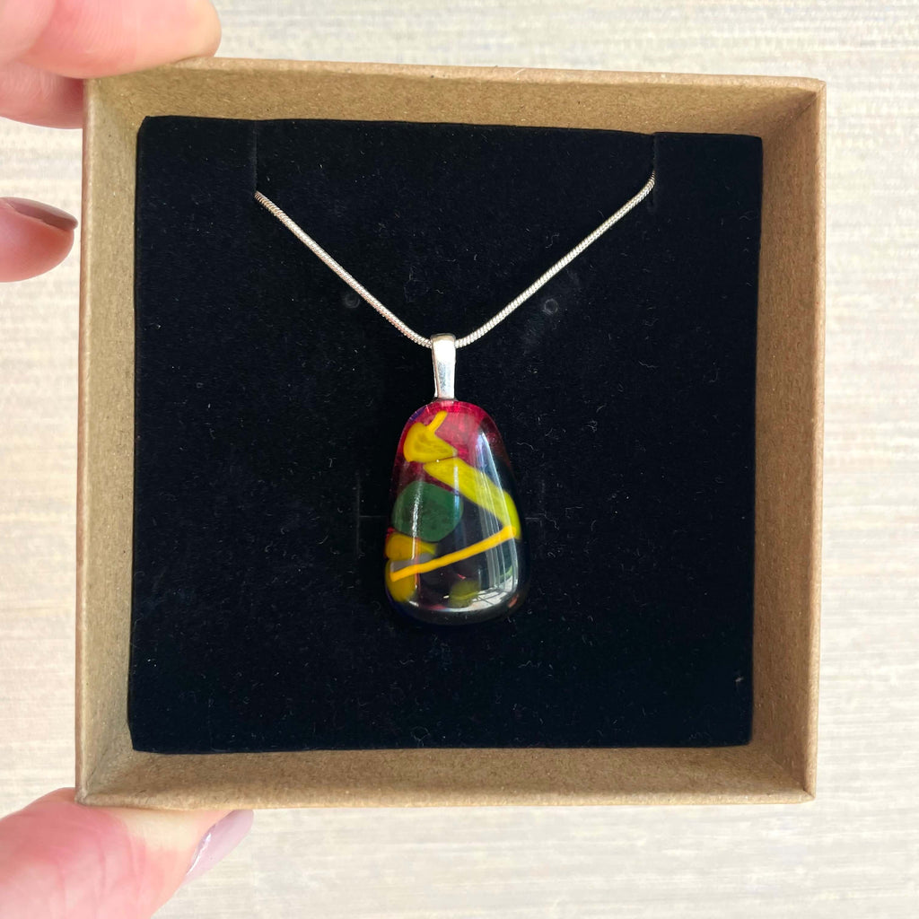 hand-crafted fused glass pendant. colourful fused glass, rich cherry red, yellow, mustard, green. on sterling silver snake chain, complete one-off, only one available, hand holing a jewellery box with pendant inside.