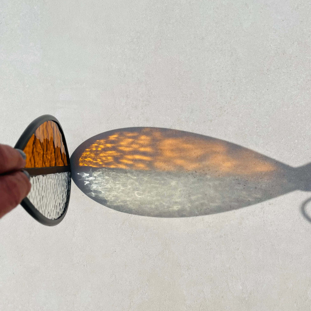 Hand holding a Stained glass medium  orange and clear glass sun catcher on a light gray background, lovely sun refraction
