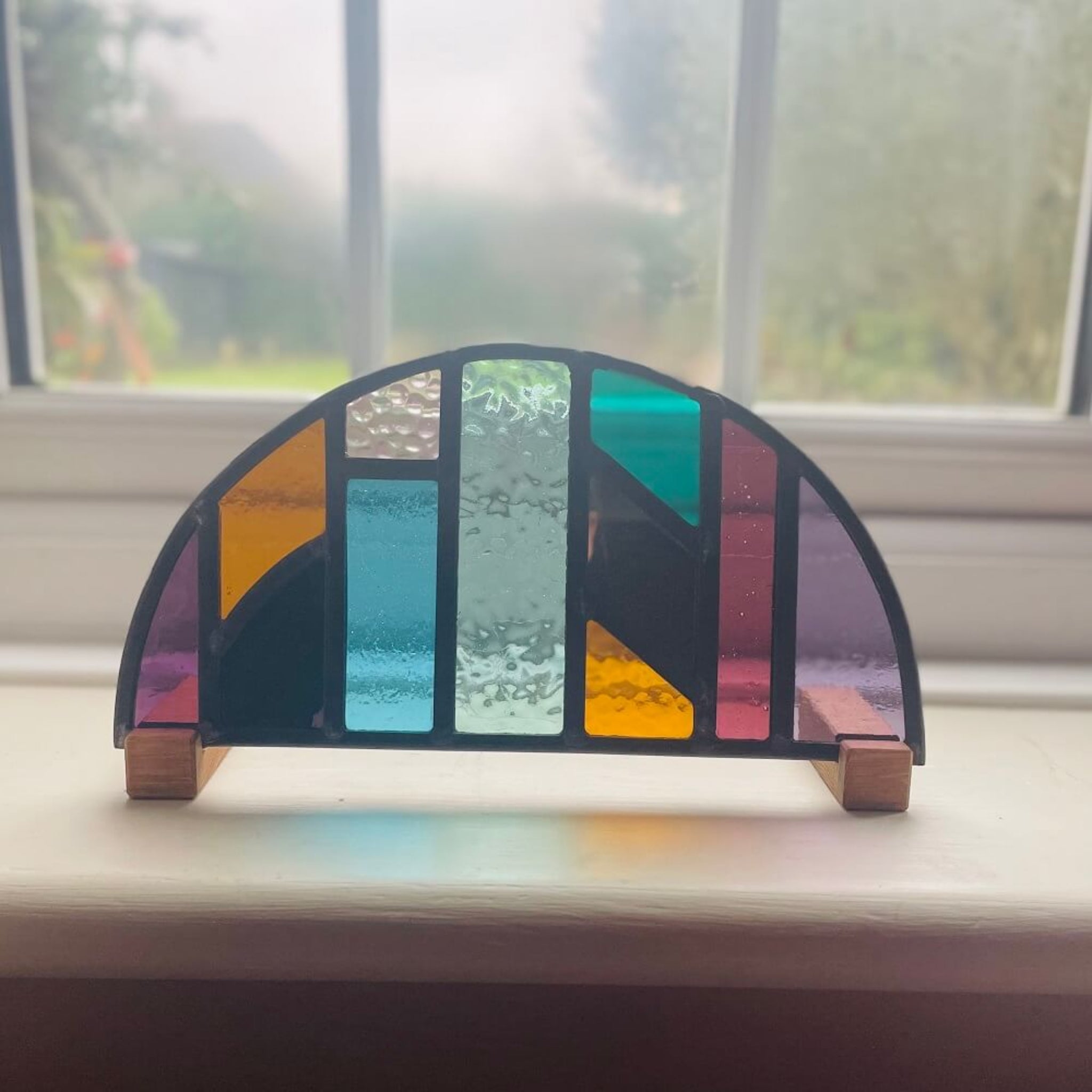 Colourful stained glass panel on a windowsill with a blurred outdoor background, Colourful arched half-circle stained glass art panel, windowsill panel, geometric shapes of coloured glass, purple, violet, orange, blue, green, mirror, . displayed on oak wooden feet, 