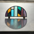 Colourful arched half-circle stained glass art panel, windowsill panel, geometric shapes of coloured glass, purple, violet, orange, blue, green, mirror, . displayed on oak wooden feet, on a stone table