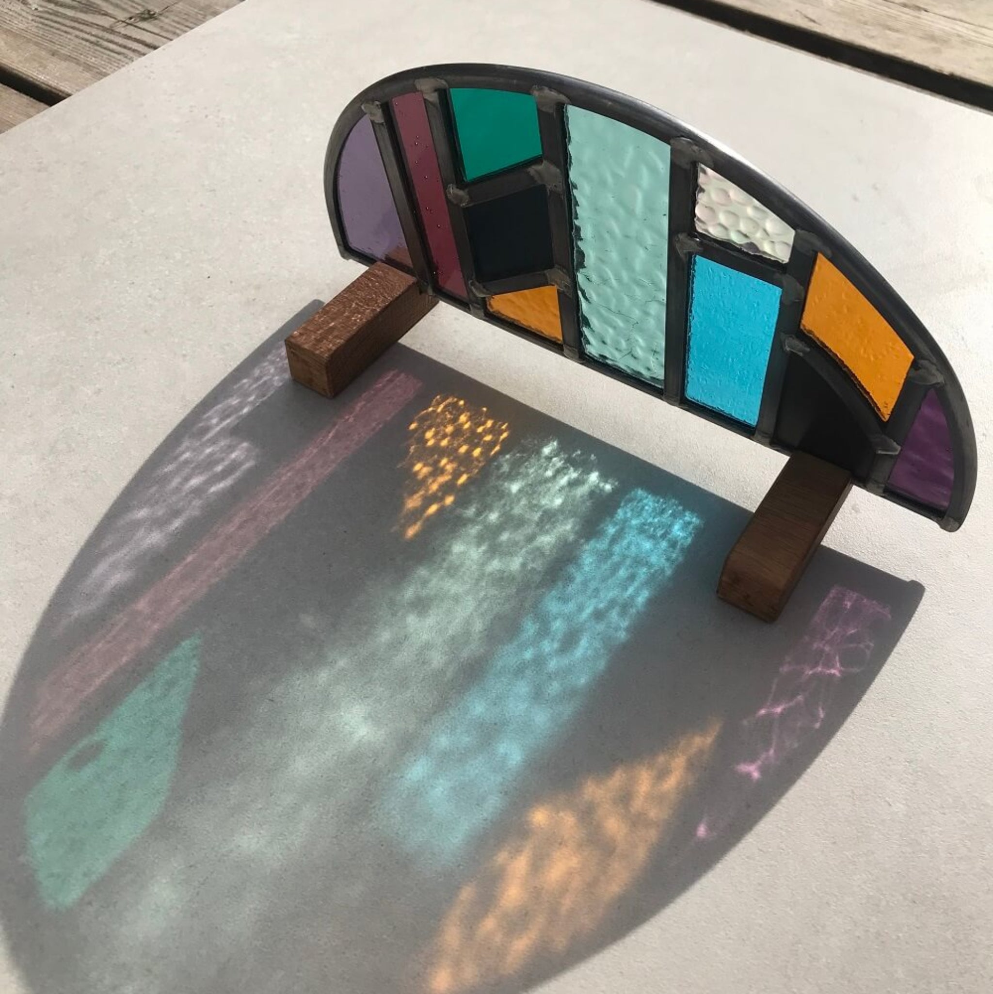 Colourful arched half-circle stained glass art panel, windowsill panel, geometric shapes of coloured glass, purple, violet, orange, blue, green, mirror,  displayed on oak wooden feet, on a stone table, with sunlight refracting onto the table creating beautiful colours on the stone table.