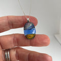 Hand holding a fused glass dichroic pendant on sterling silver fine chain, gold, blue and purple textured glittering glass, against a white background