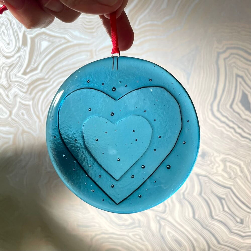 fused glass 'love bomb' Blue heart-shaped hanging decoration, ornament with a red string on a patterned creamy white background