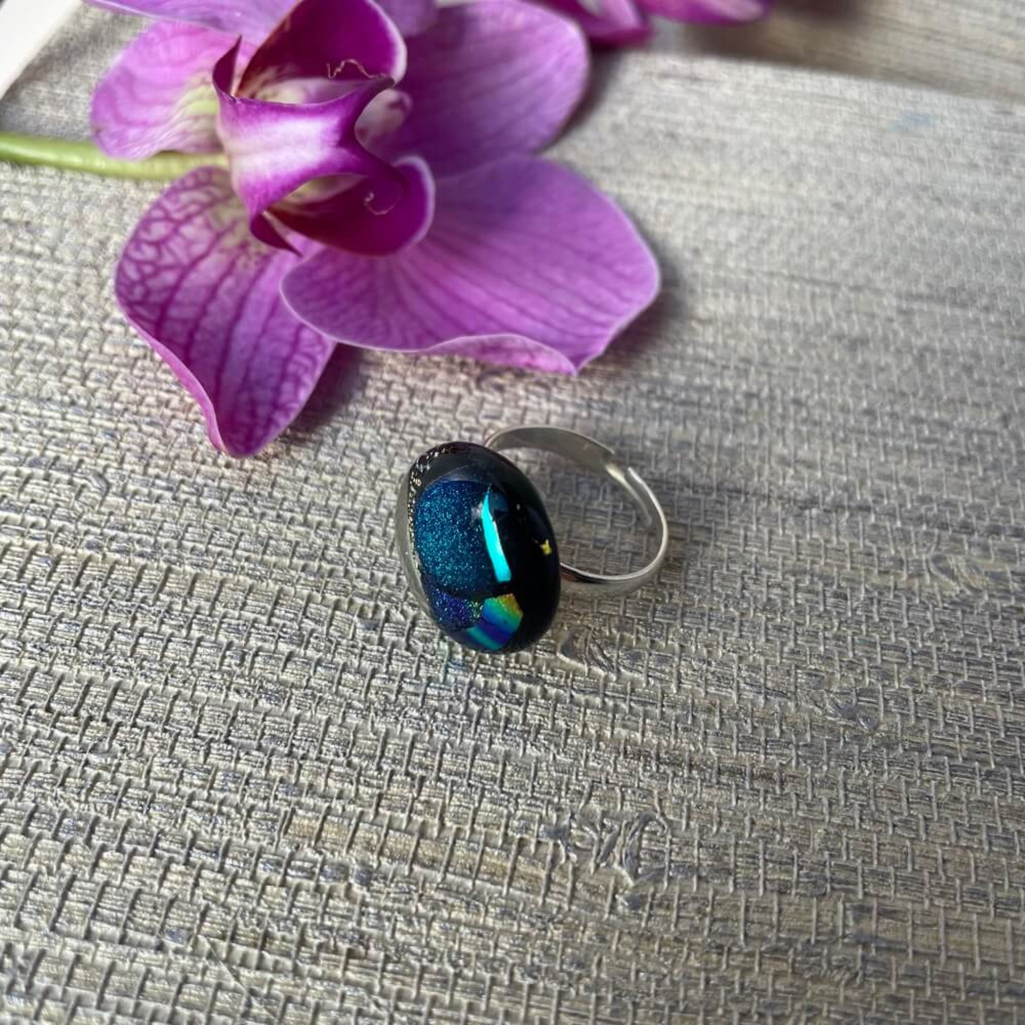 Close-up of a round jewel statement ring, colourful dichroic fused glass ring, yellow, blue, glittering pink glass, with purple orchids in the background, on adjustable stainless steel ring base