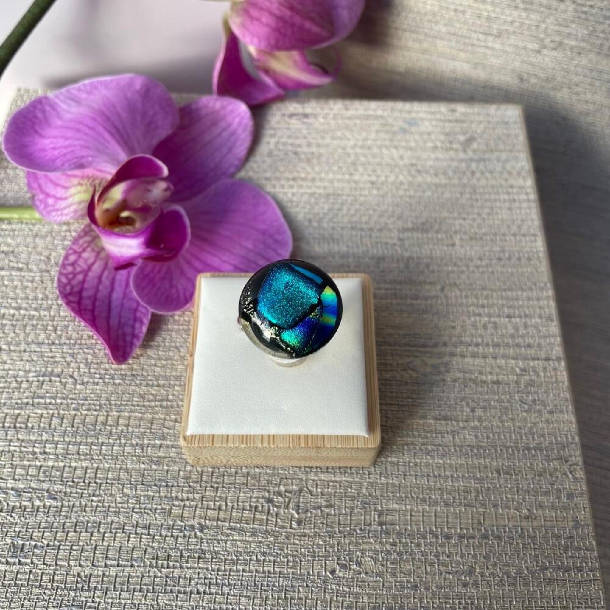 Close-up of a round jewel statement ring, colourful dichroic fused glass ring, yellow, blue, glittering pink glass, with purple orchids in the background, on adjustable stainless steel ring base