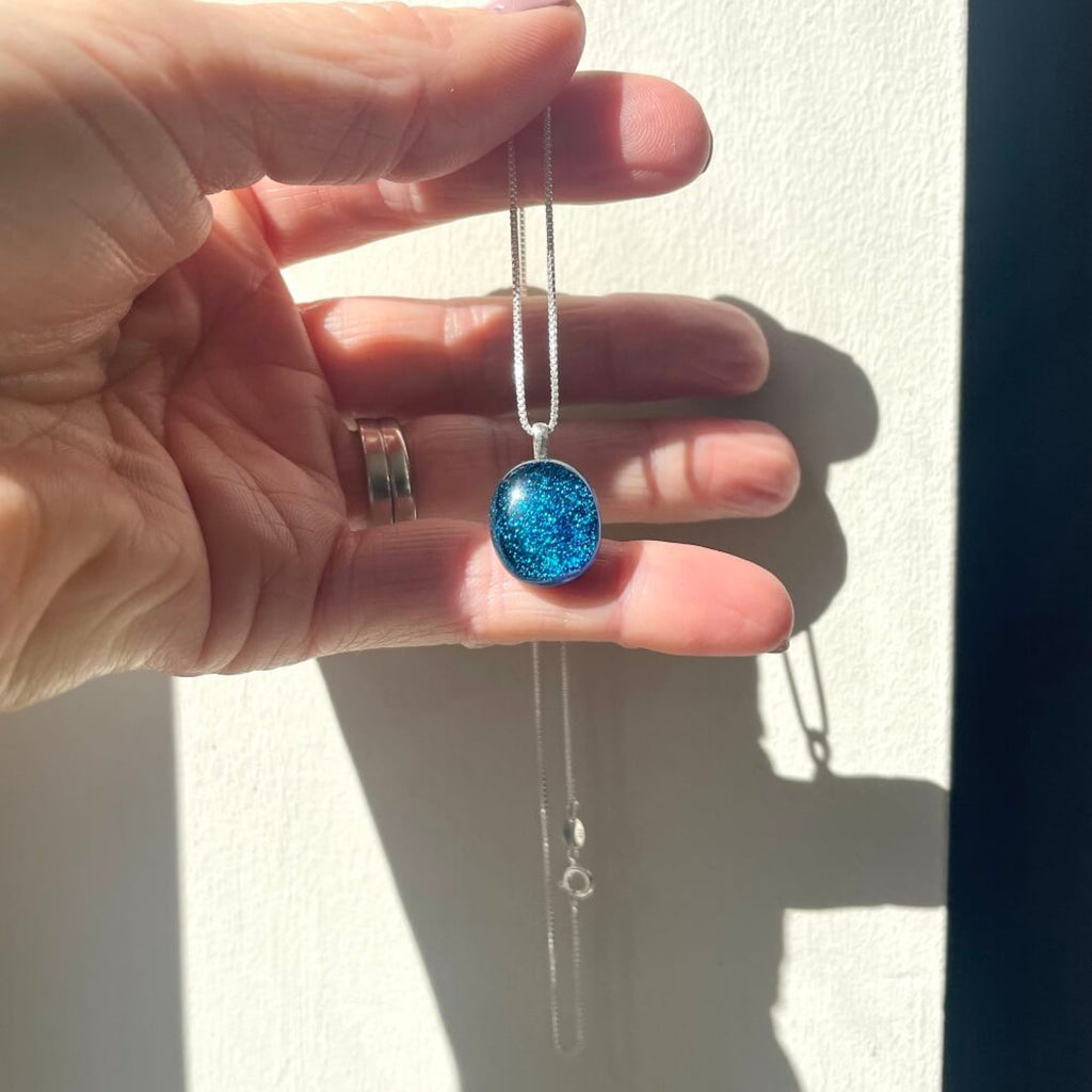 Hand holding a handmade necklace with a fused glass blue dichroic shimmering  pendant, on a sterling silver elegant box chain, against a neutral background