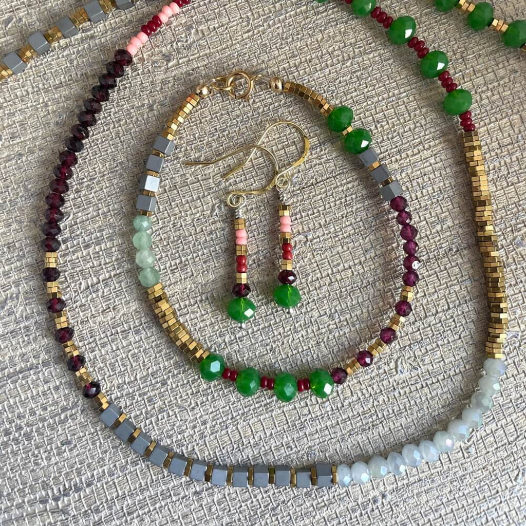 Beaded jewellery set with necklace, bracelet, and earrings on a textured surface, colourful beads, red faceted garnets, golden hematite, silver cubed hematite, 14k rolled gold finding and gold vermeil earring hooks