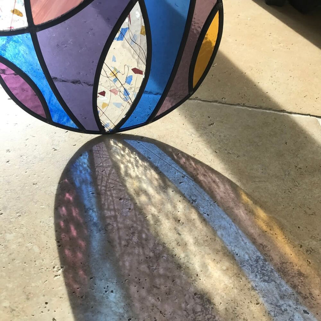 Large stained glass art panel, hanging panel, colourful curved shapes of stained glass, purple, violet, blue, yellow glass,  sunlight refracting through the glass onto a stone floor