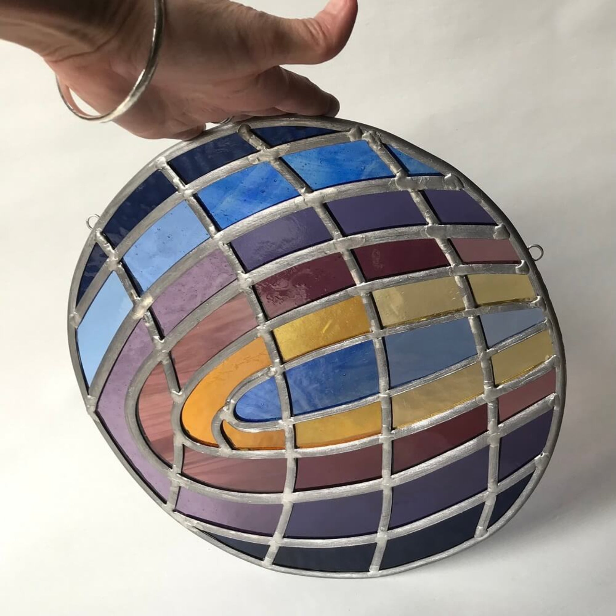 Large disk shaped, round stained glass art panel with multi colored curved shapes, purple, blue, violet, yellow tones of stained glass, held by a hand against a white background