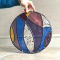 Large stained glass art panel, hanging panel, colourful curved shapes of stained glass, purple, violet, blue, yellow glass,  held by a hand against a neutral background