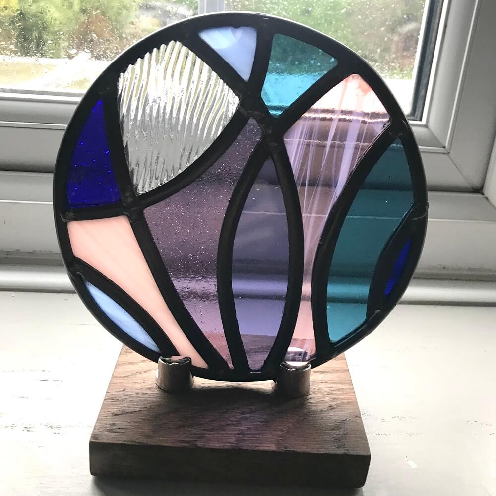 stained glass disk panel artwork, curved shapes of glass, blue and purples, Stained glass piece on a wooden stand with a window in the background