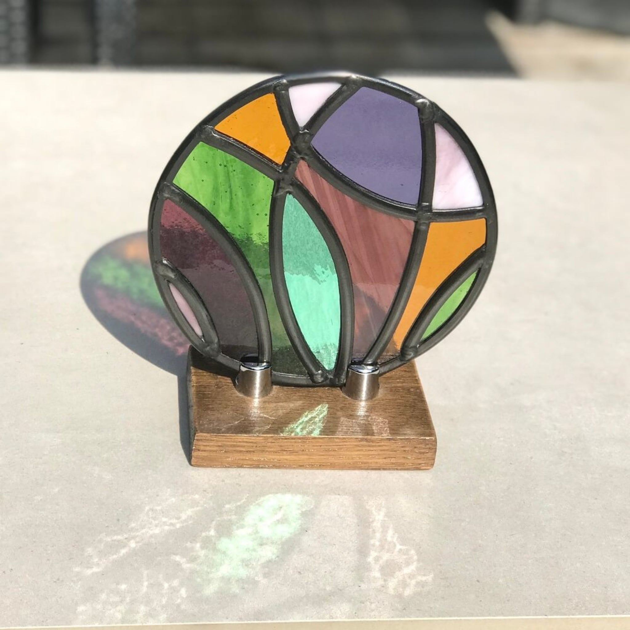  a colorful stained glass artwork disk panel, in the sun, with purple, pink, violet, green, yellow glass,  curved disk design on a wooden base, on a stone table