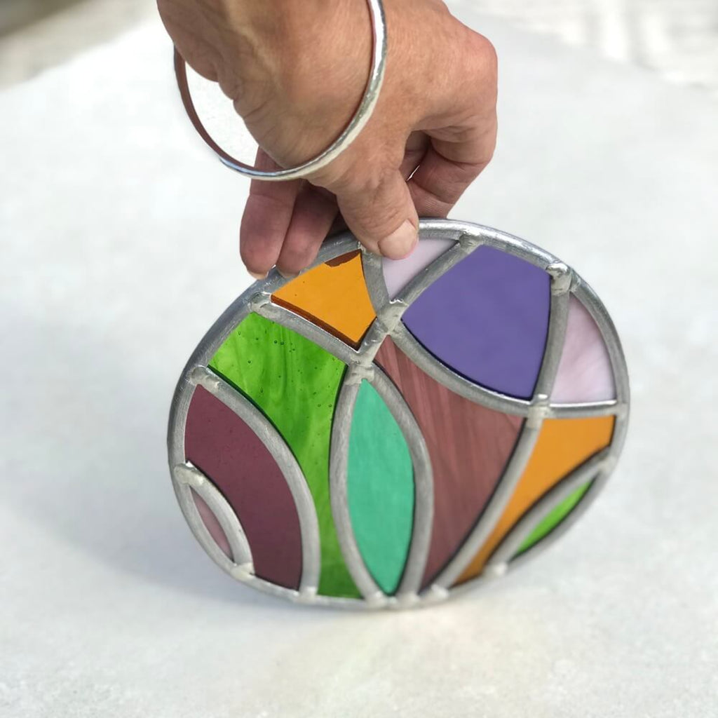 Hand holding a colorful stained glass artwork disk panel, with purple, pink, violet, green, yellow glass, on a white background