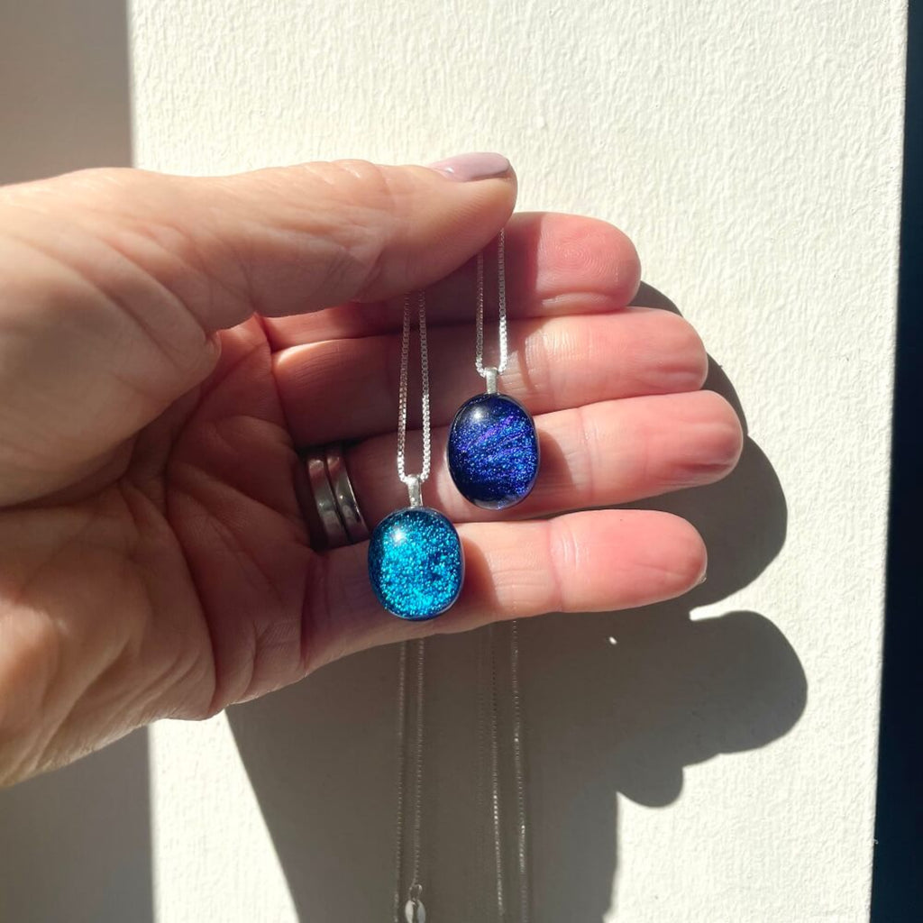 Hand holding 2 handmade necklaces, fused glass blue dichroic shimmering  pendant, and a purple pendant, both on sterling silver elegant box chains, against a neutral back