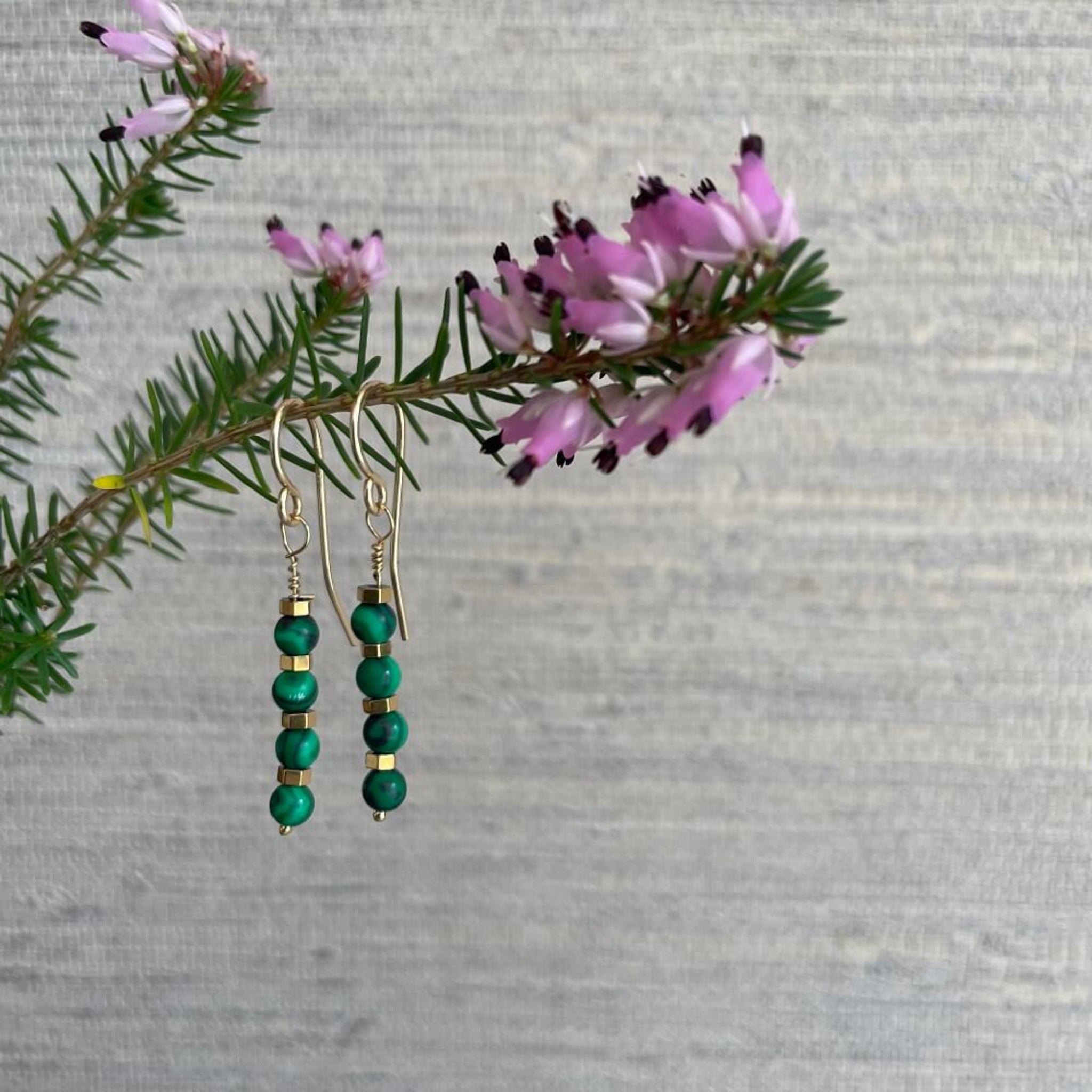 Green beaded malachite and 14k rolled gold, gold hematite beads earrings on a branch with pink flowers against a gray background