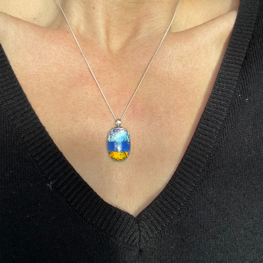 H fused glass dichroic pendant on sterling silver fine chain, gold, blue and purple textured glittering glass, colorful pendant against a women's chest black v neck