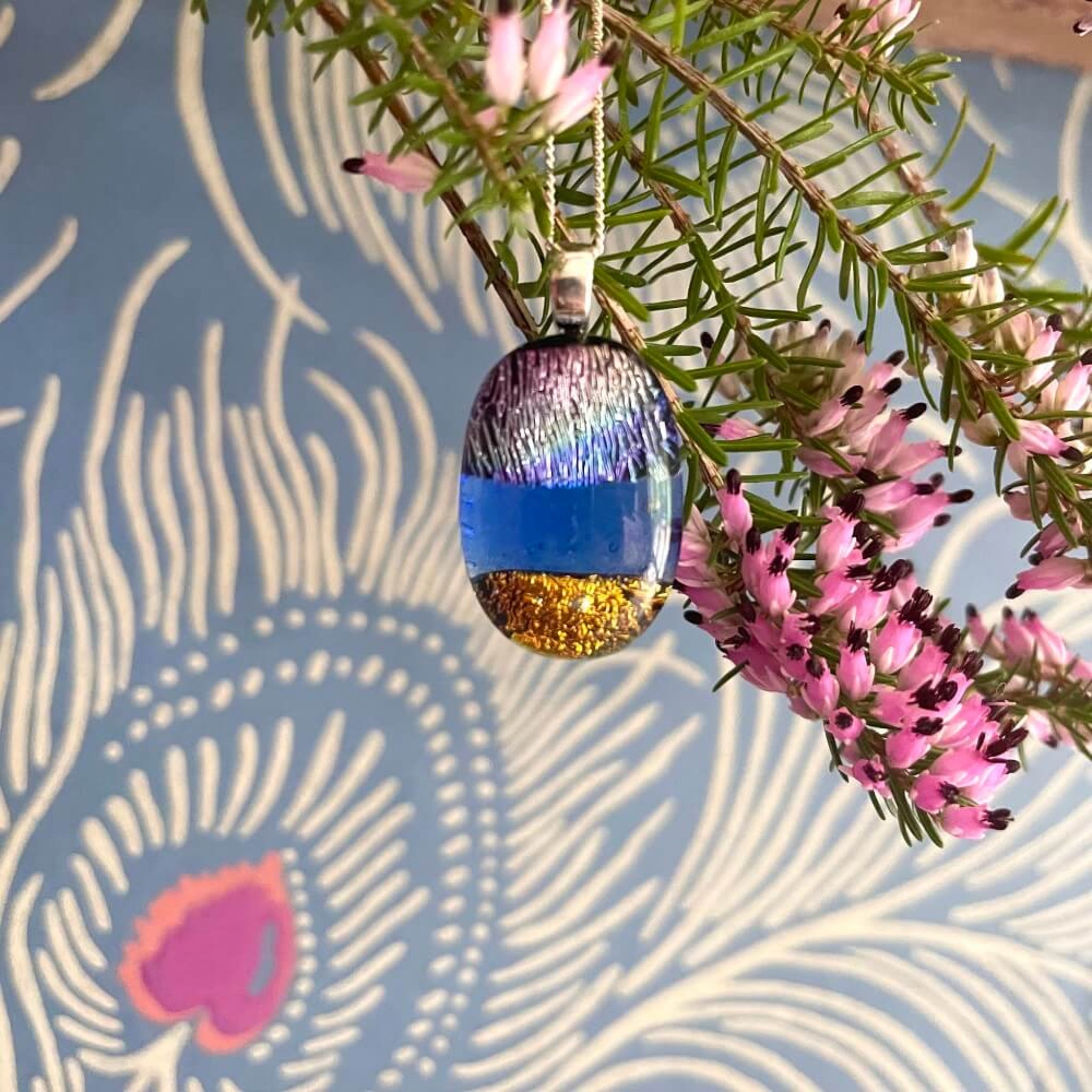  fused glass dichroic pendant on sterling silver fine chain, gold, blue and purple textured glittering glass, background, hanging from a branch with pink flowers against a patterned background