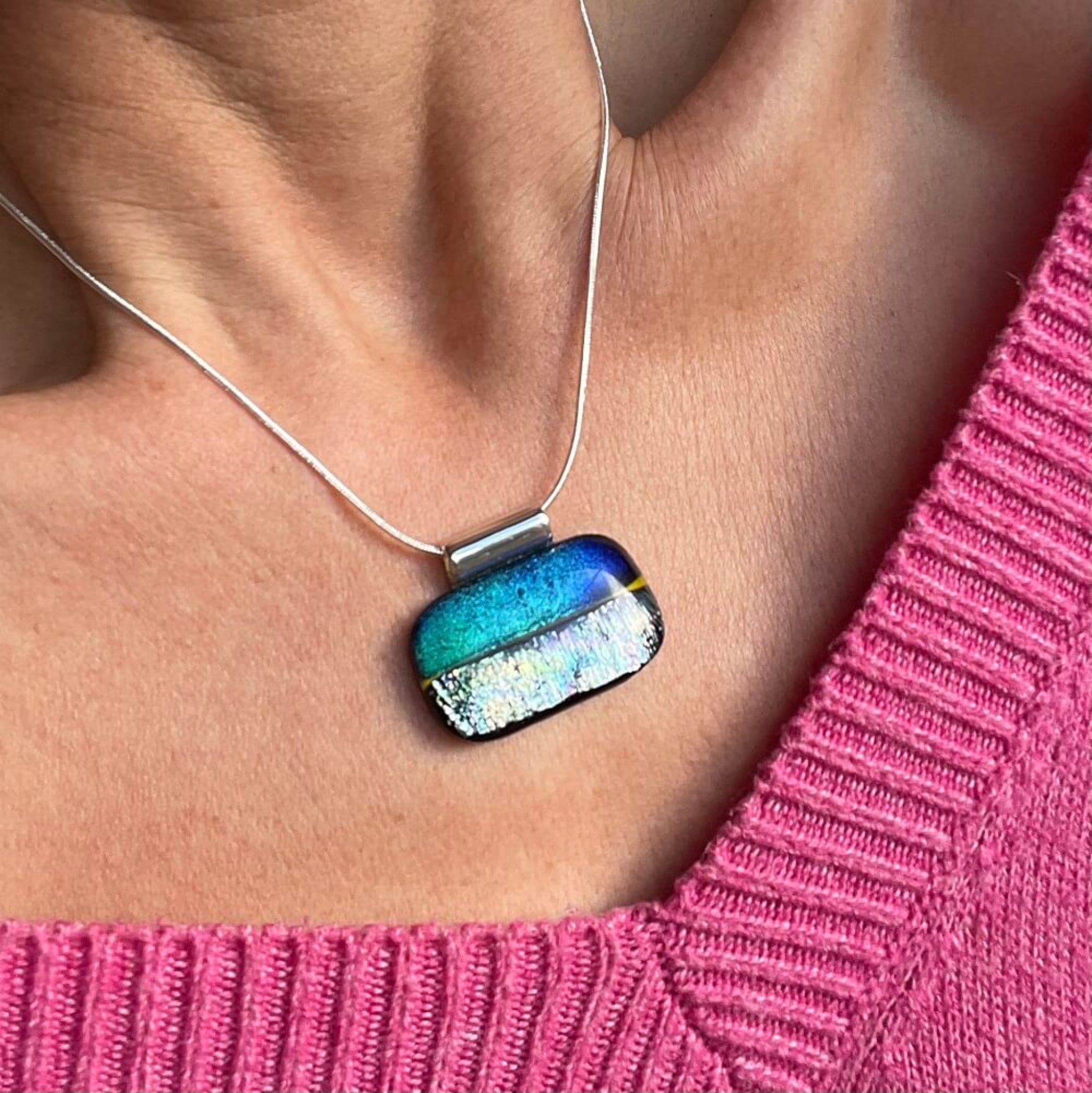 colorful pendant worn by a person in a pink sweater. shimmering blue, yellow and silver dichroic glass, rectangle statement pendant, on sterling silver snake chain, stainless steel bail, 