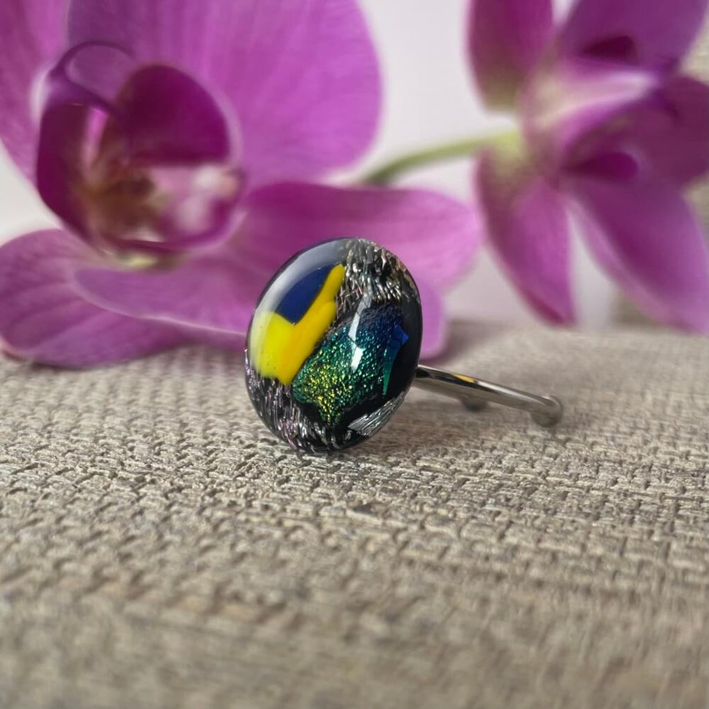 Close-up of a round jewel statement ring, colourful dichroic fused glass ring, yellow, blue, glittering pink glass, with purple orchids in the background