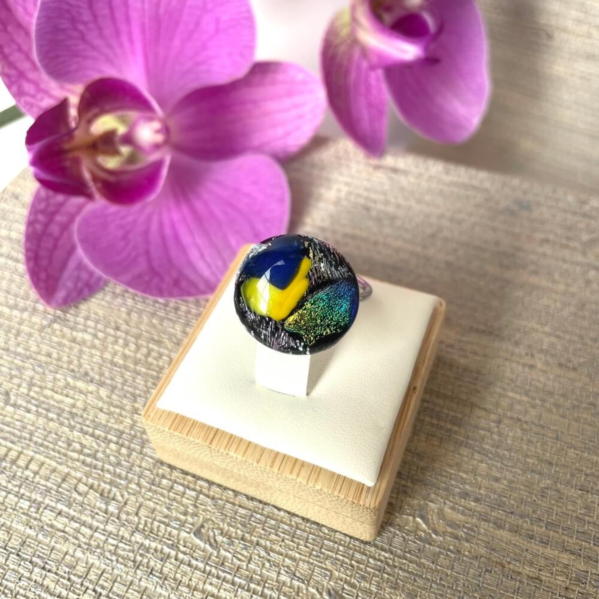 Close-up of a round jewel statement ring, colourful dichroic fused glass ring, yellow, blue, glittering pink glass, with purple orchids in the background