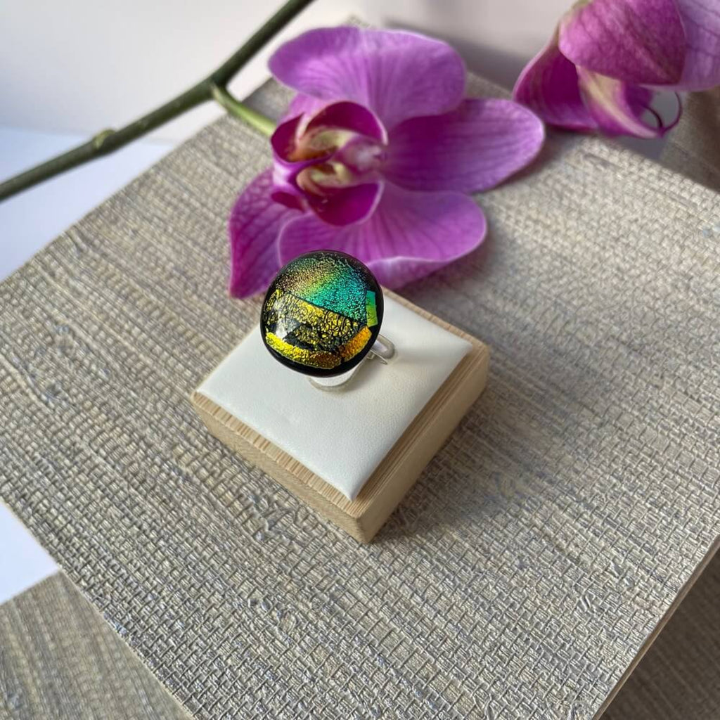 Close-up of a round jewel statement ring, colourful dichroic fused glass ring, yellow, blue, glittering pink glass, with purple orchids in the background, on adjustable stainless steel ring base, pink orchid in background
