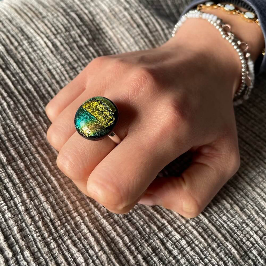 hand with fused glass Ring, Close-up of a round jewel statement ring, colourful dichroic fused glass ring, yellow, blue, glittering pink glass, with purple orchids in the background, on adjustable stainless steel ring base