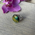 Close-up of a round jewel statement ring, colourful dichroic fused glass ring, yellow, blue, glittering pink glass, with purple orchids in the background, on adjustable stainless steel ring base