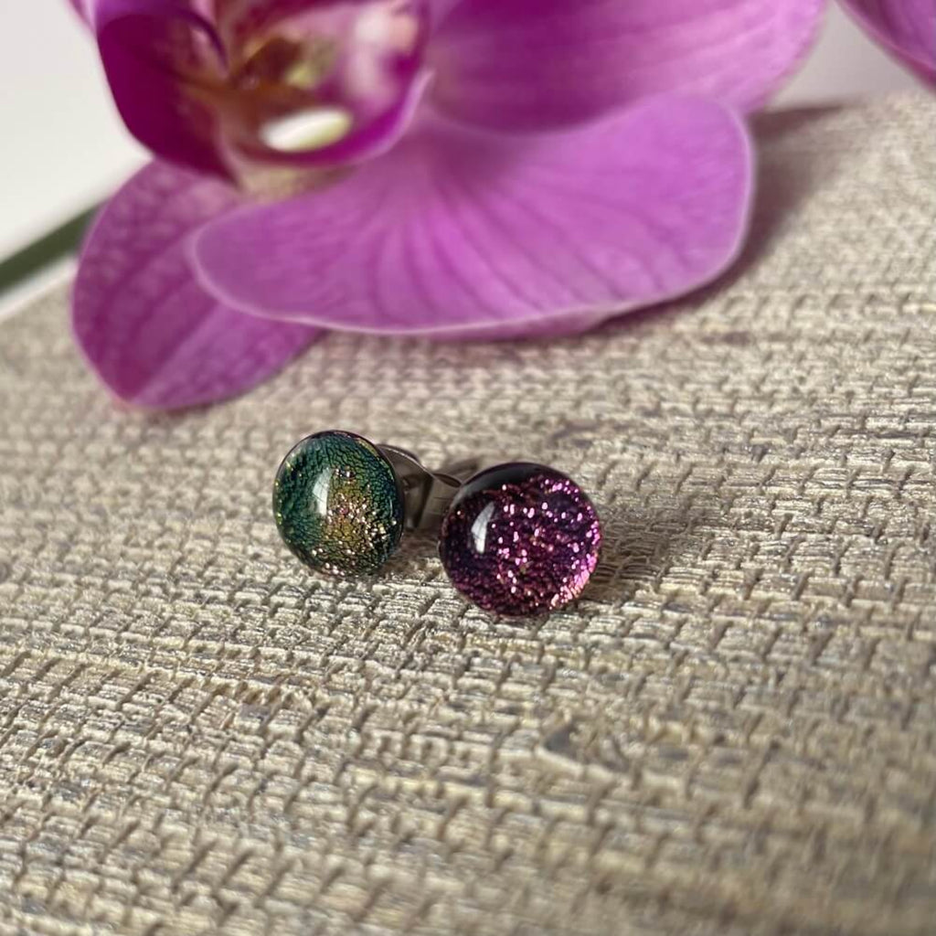 Pair of round fused glass earrings, glittering dichroic fused glass, with glittery finish on a textured surface with a purple flower in the background.