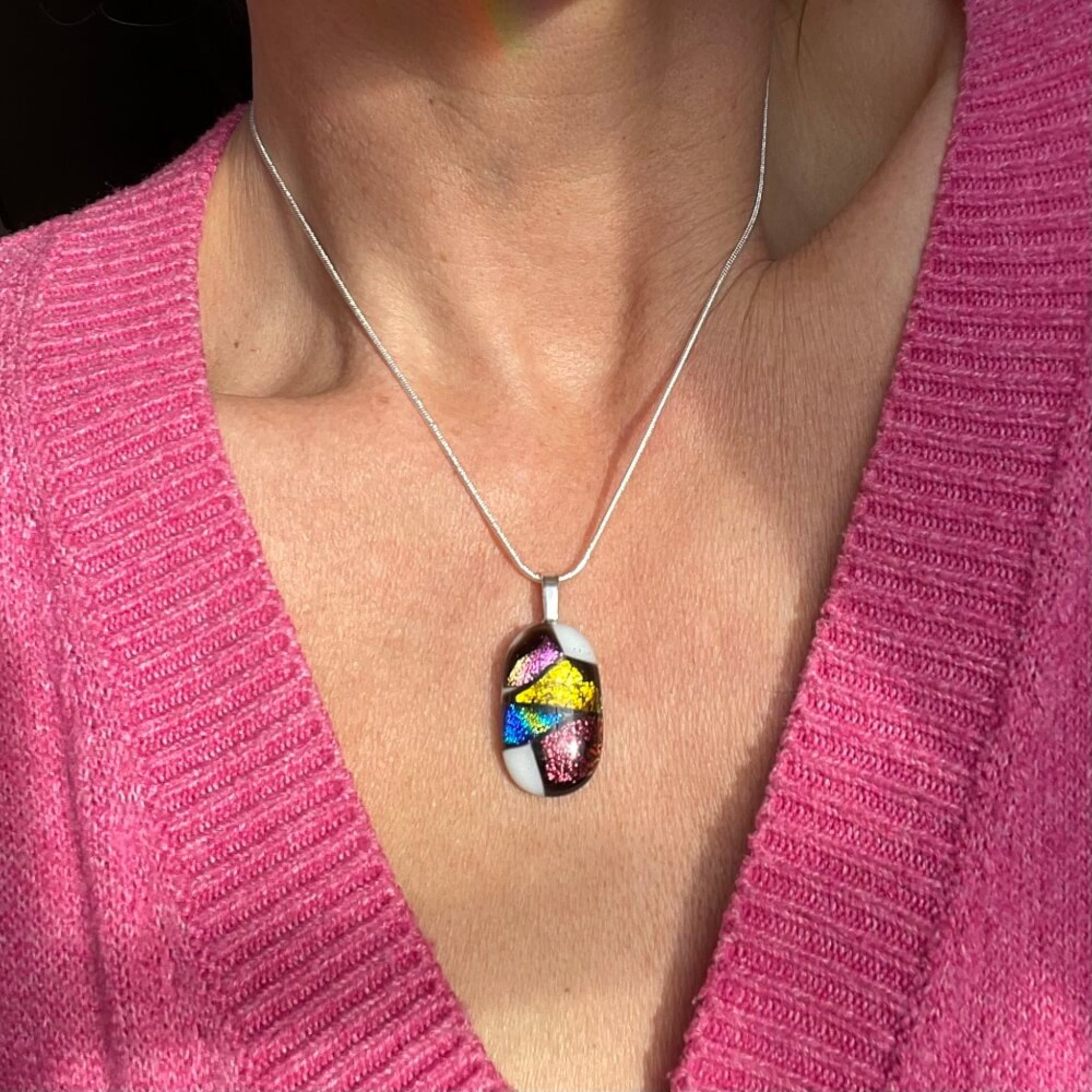 fused glass pedant, colourful pendant worn by a person in a pink sweater. pink, gold, blue, purple glittering dichroic glass, in a sterling silver snake chain.