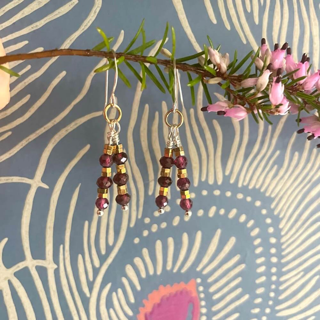 Beaded red wine garnet and gold hematite beaded dangly earrings, with 14k rolled gold and sterling silver earring hooks. hanging from a branch with pink flowers on a patterned background