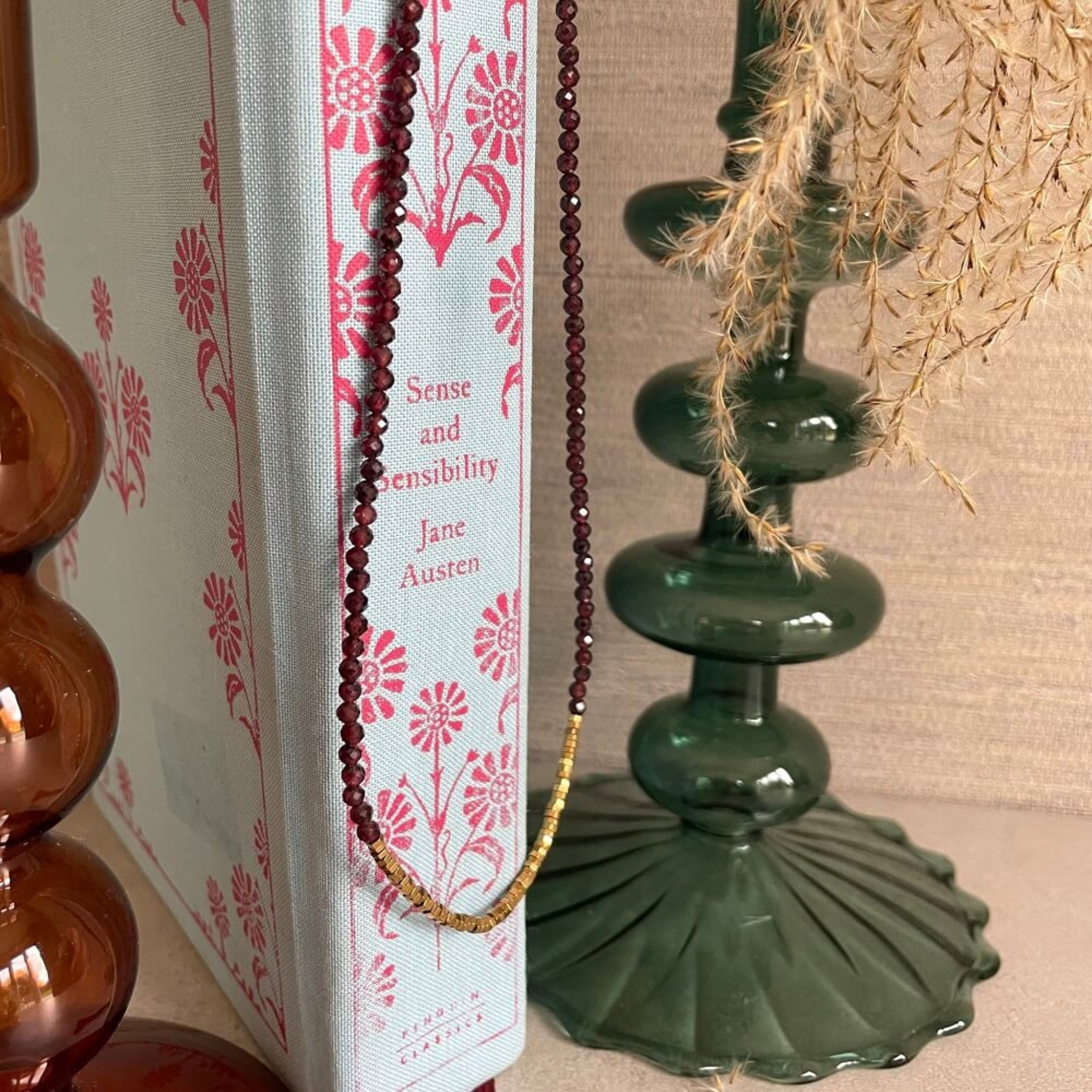 Book titled 'Sense and Sensibility' by Jane Austen with decorative garnet and hematite beads on a floral-patterned background, necklace handmade with 14k rolled gold fittings,