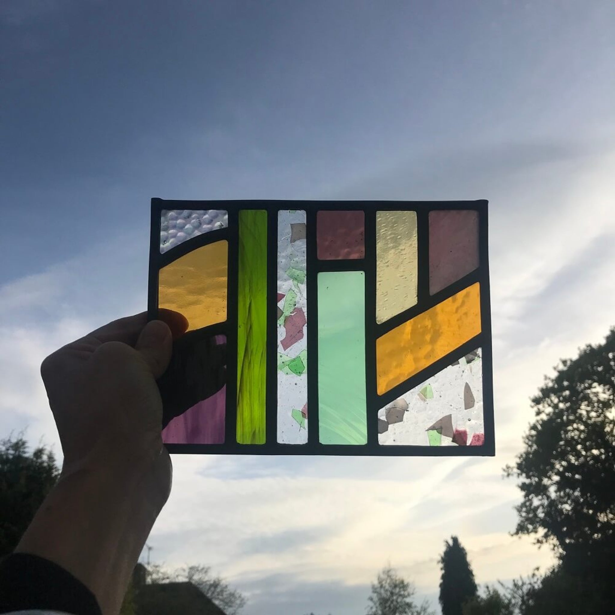 Hand holding a colourful geometric stained glass panel against a blue sky, with trees. stained glass art panel, windowsill panel, geometric shapes of coloured glass, purple, violet, orange, blue, green, 