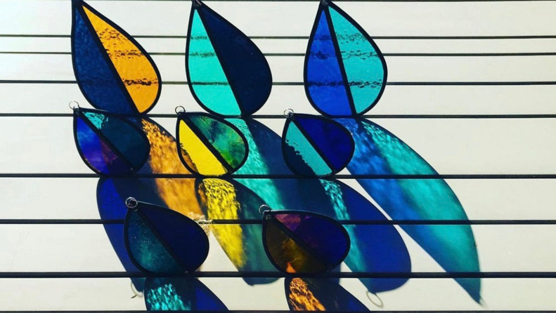 set of colorful stained glass leaf design garden 'tree spinners' / window 'sun catchers' arranged on a table with sunlight beaming through them, creating lovely colourful refraction's, or orange, blue, yellow, aqua stained glass colours