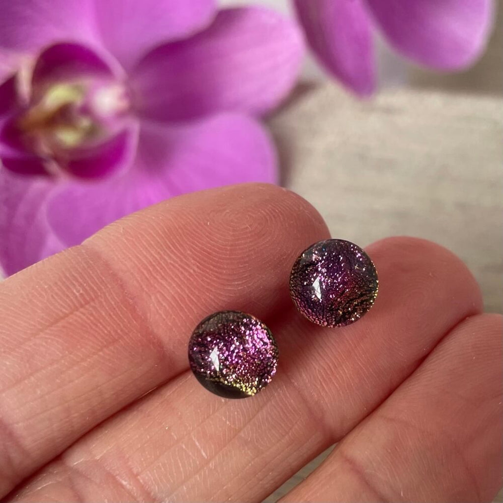 Purple / pink / green glittery fused glass earrings, dichroic glass, held between fingers with a blurred purple flower in the background