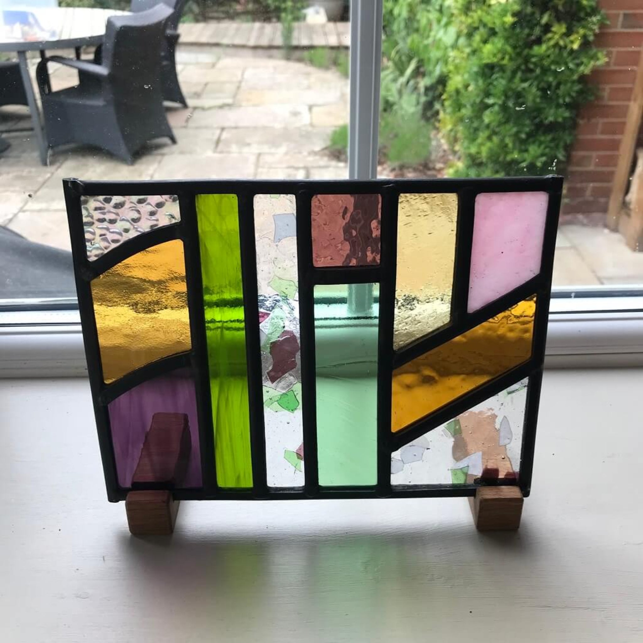 Colourful geometric stained glass art piece on a stand with a garden view in the background,  stained glass art panel, windowsill panel, geometric shapes of coloured glass, purple, violet, orange, blue, green, displayed on oak wooden feet, 