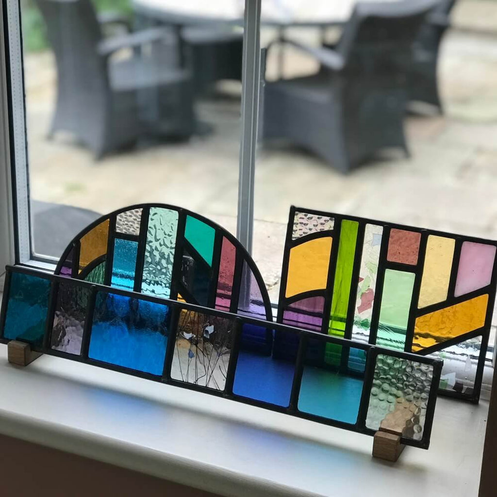 set of 3 colourful geometric stained glass art panels on a windowsill, Colourful arched half-circle stained glass art panel, windowsill panel, geometric shapes of coloured glass, purple, violet, orange, blue, green, mirror, . displayed on oak wooden feet,