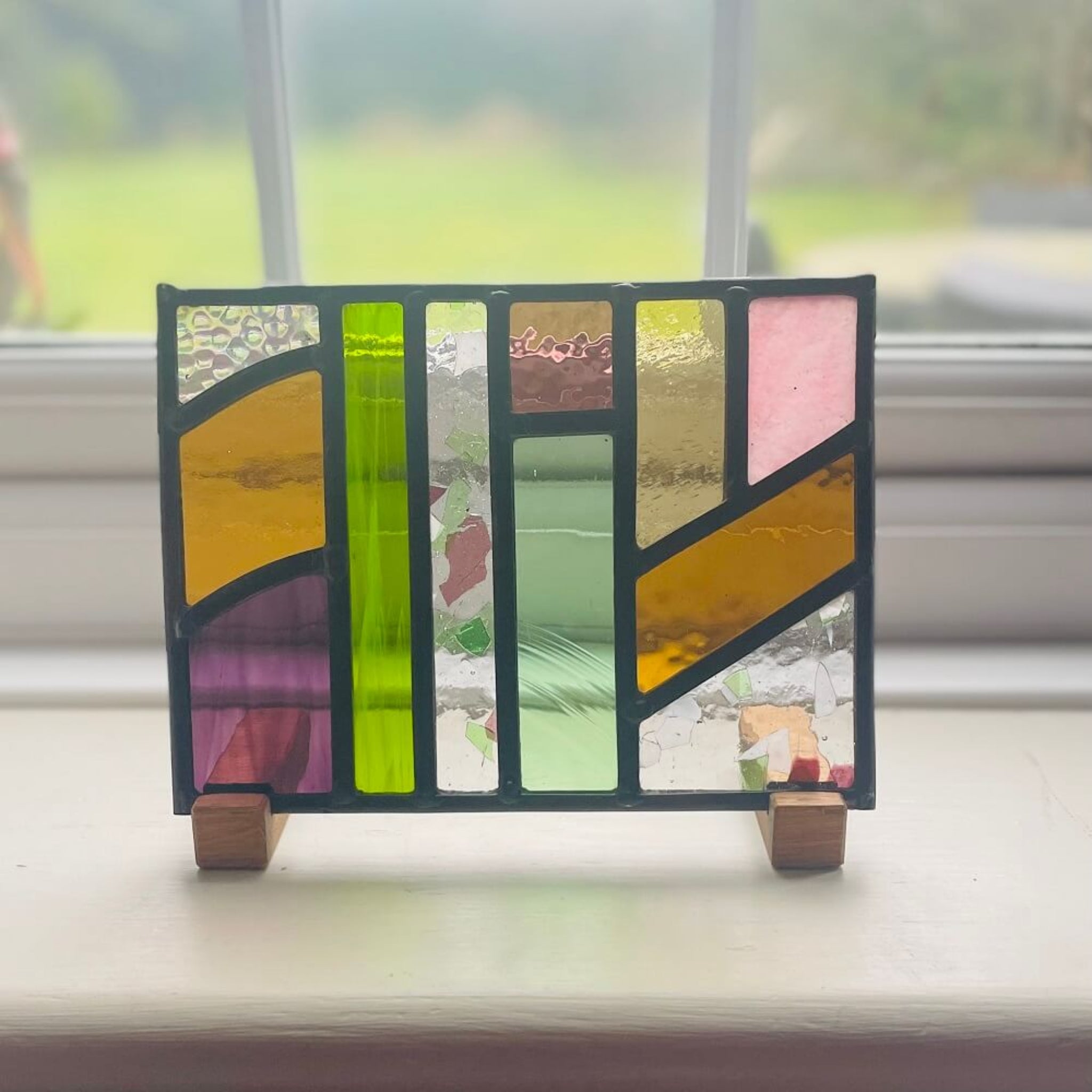 Colourful stained glass art panel, windowsill panel, geometric shapes of coloured glass, purple, violet, orange, blue, green,  displayed on oak wooden feet, on a windowsill