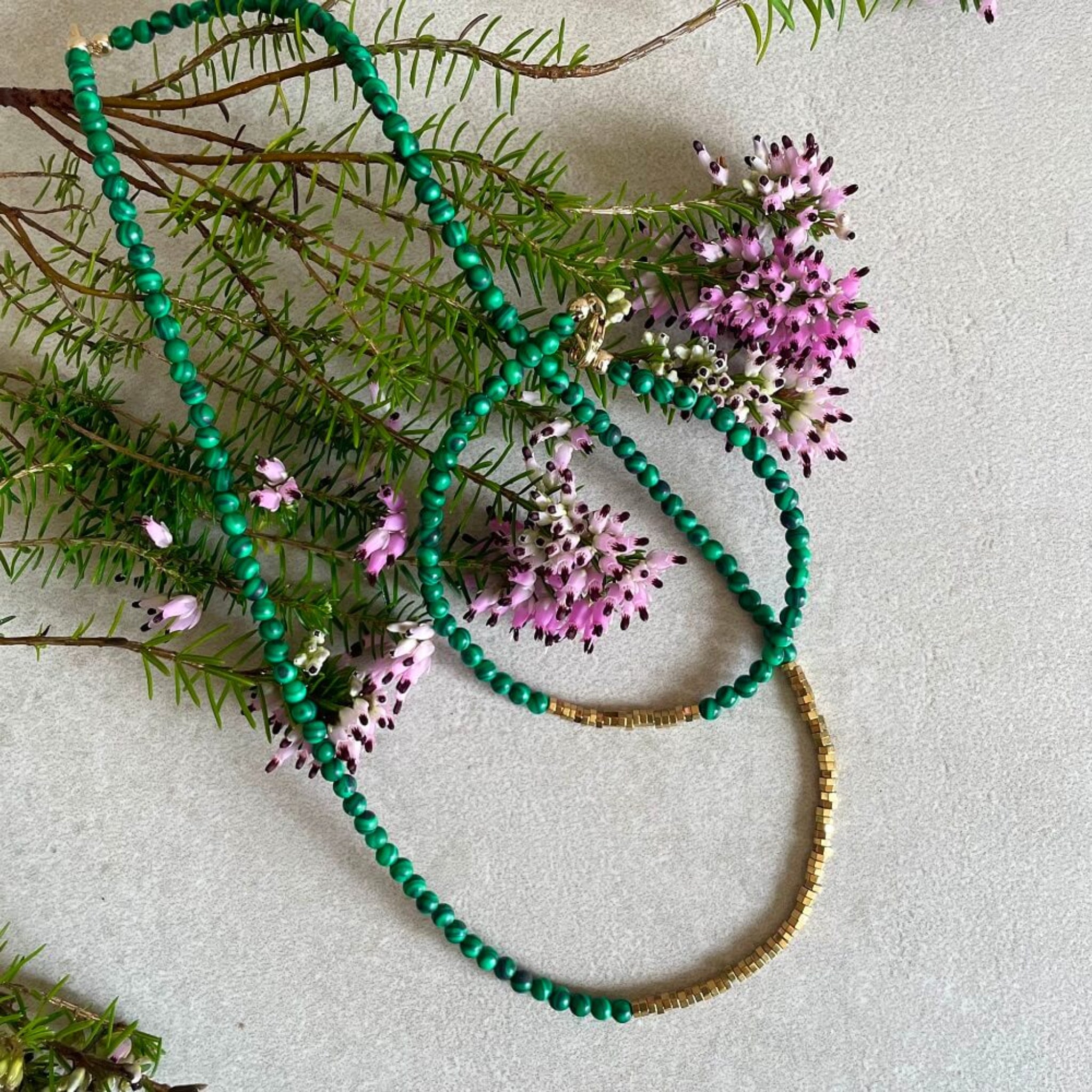 Green beaded malachite necklace and bracelet with gold hematite accents on a light gray background with pink flowers and green leaves, 14k rolled gold findings