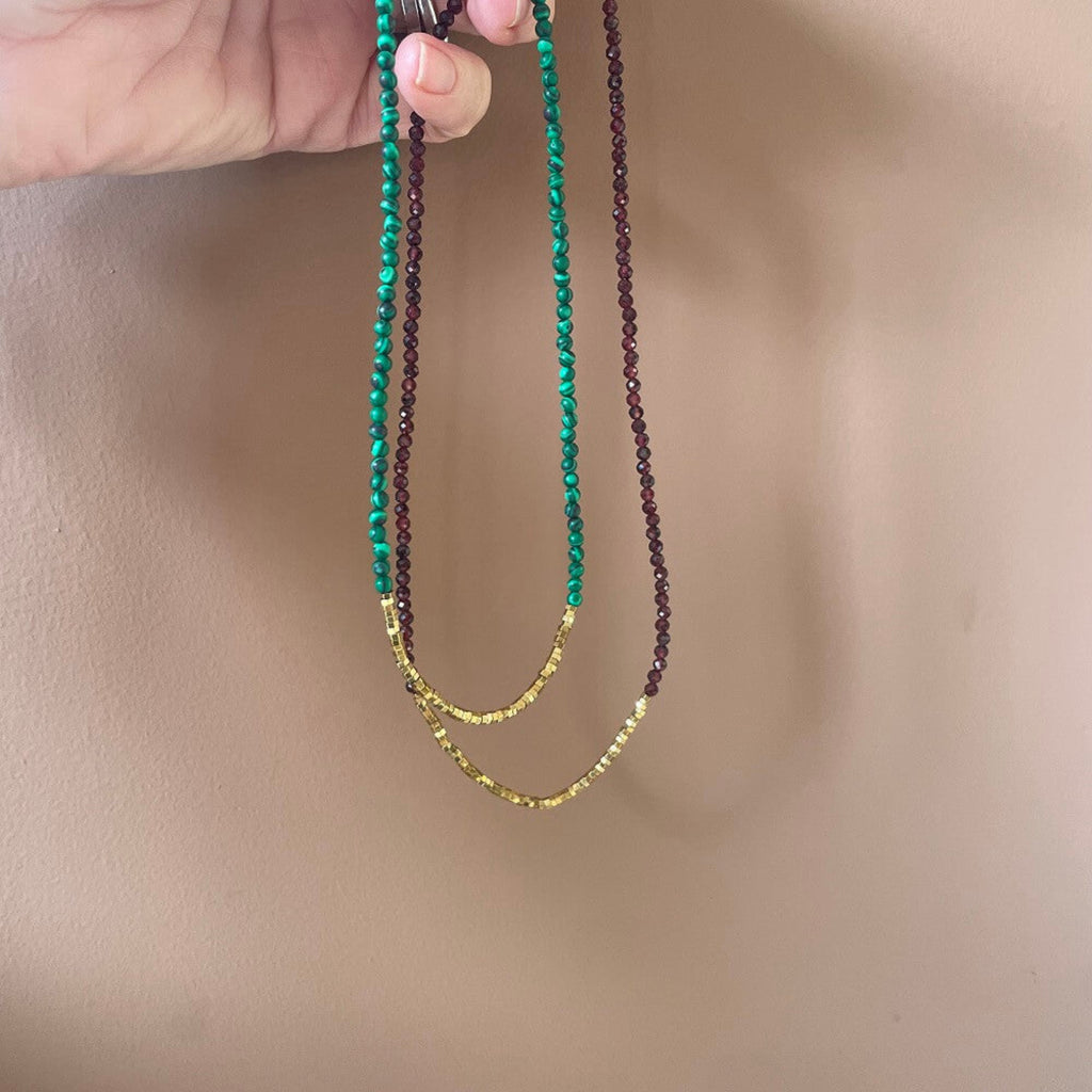 Two beaded necklaces, one green malachite and gold hematite, and one ruby red faceted garnet and gold hematite, made with quality 14k rolled gold fittings,  held against a terracotta background