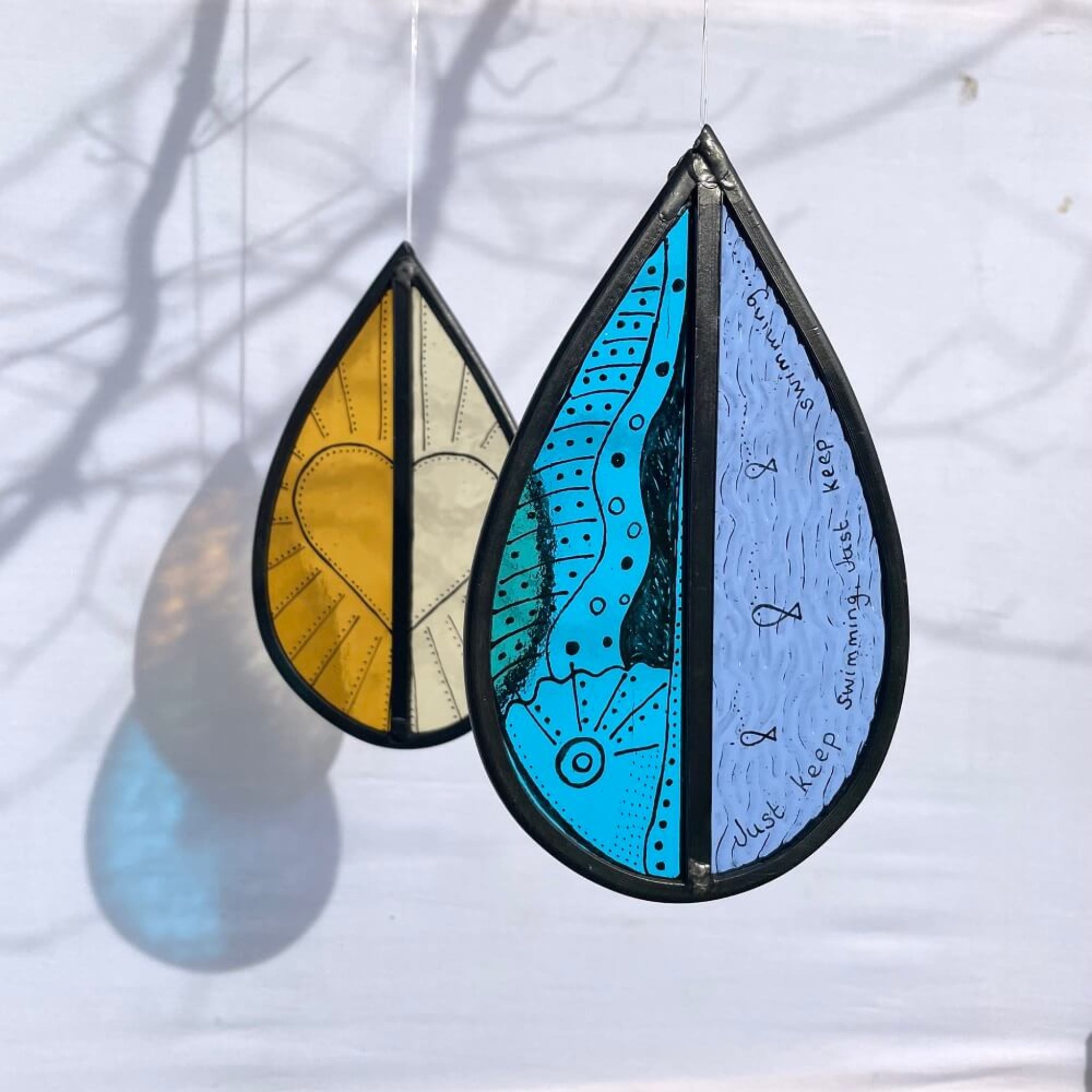 Two teardrop-shaped stained glass hanging tree spinners / window sun catcher, with hand painted fish, and words 'just keep swimming' and orange and yellow stained glass, with heart painted design, hanging against  a white background, 