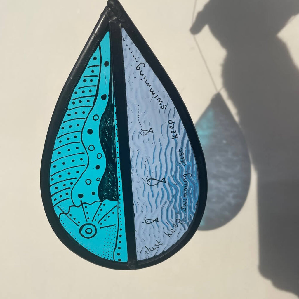 teardrop-shaped stained glass garden tree spinner /  window sun catcher, fish drawing with 'just keep swimming' text,  hanging against a white background. lovely blue sunlight refraction, 