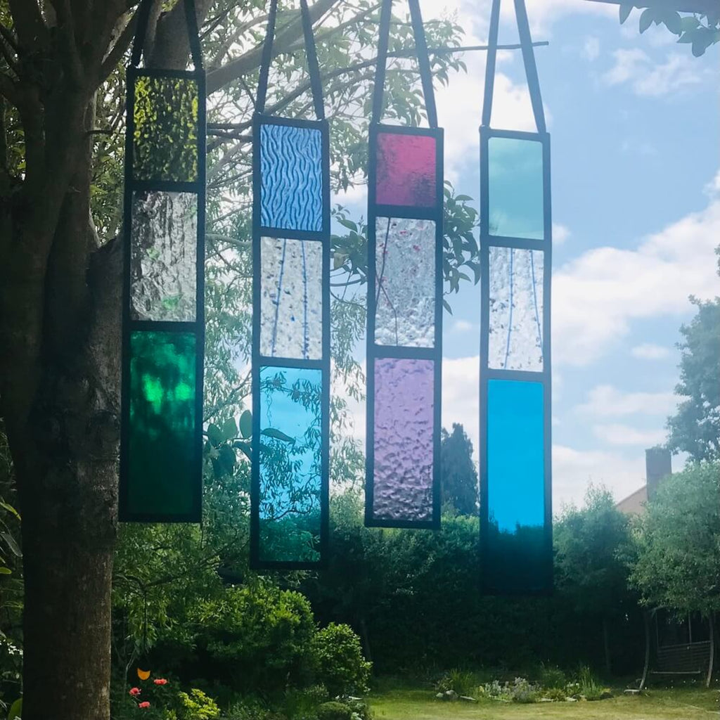 Colourful stained glass panels hanging outdoors with trees and sky in the background, rectangle stained glass panels, 3 sections of glass, blue, purple, green glass, 