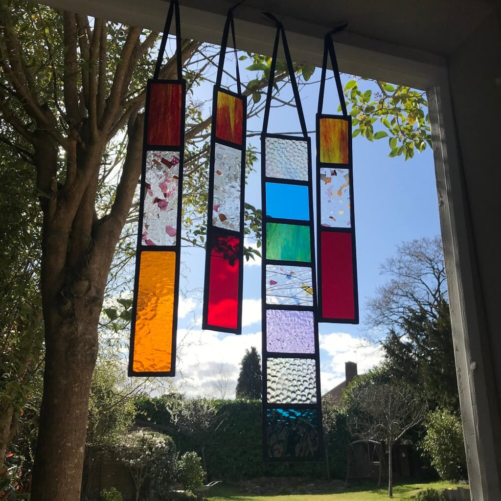 group of 4 Colourful stained glass panels hanging indoors with a garden view outside. red and orange coloured stained glass, rectangle tree spinners, or sun catchers. 