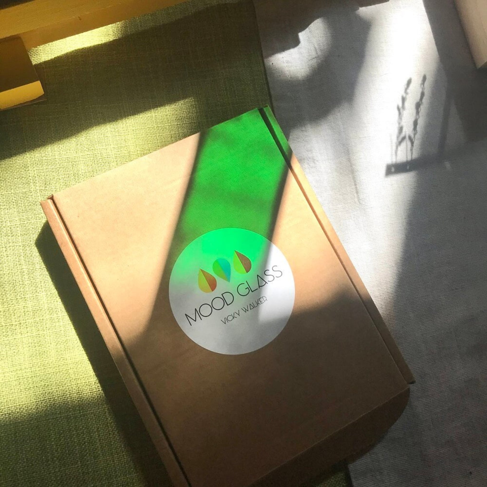 Box with a 'Mood Glass' sticker on a textured surface, green sun refraction from glass sun catcher