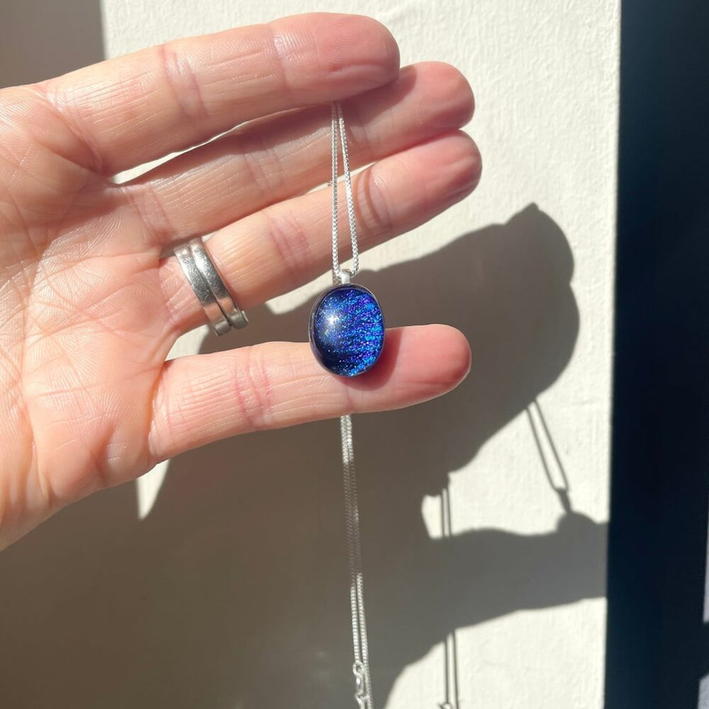 Hand holding a handmade necklace with a fused glass purple dichroic shimmering  pendant, on a sterling silver elegant box chain, against a neutral background