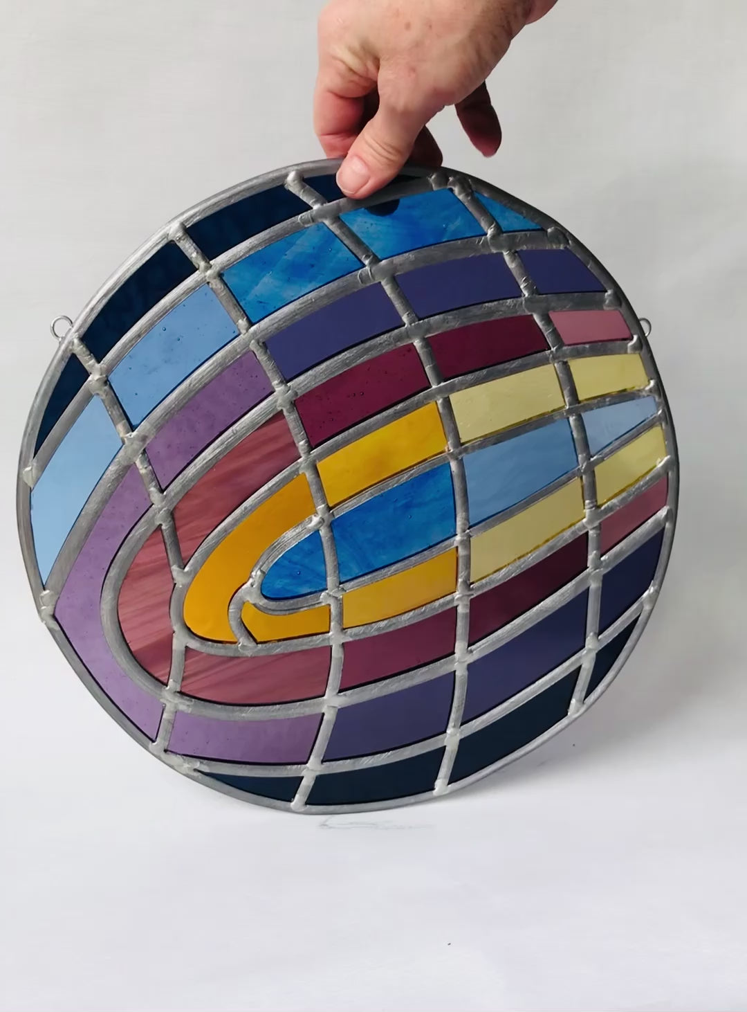 video of a hand spinning a Large round stained glass art panel / piece with colorful stained glass curved shapes, of blue, purple, gold, violet. geometric design on a white background