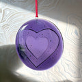 handmade fused glass Purple heart-shaped 'love bomb' hanging decoration / ornament with a red ribbon on a textured white background