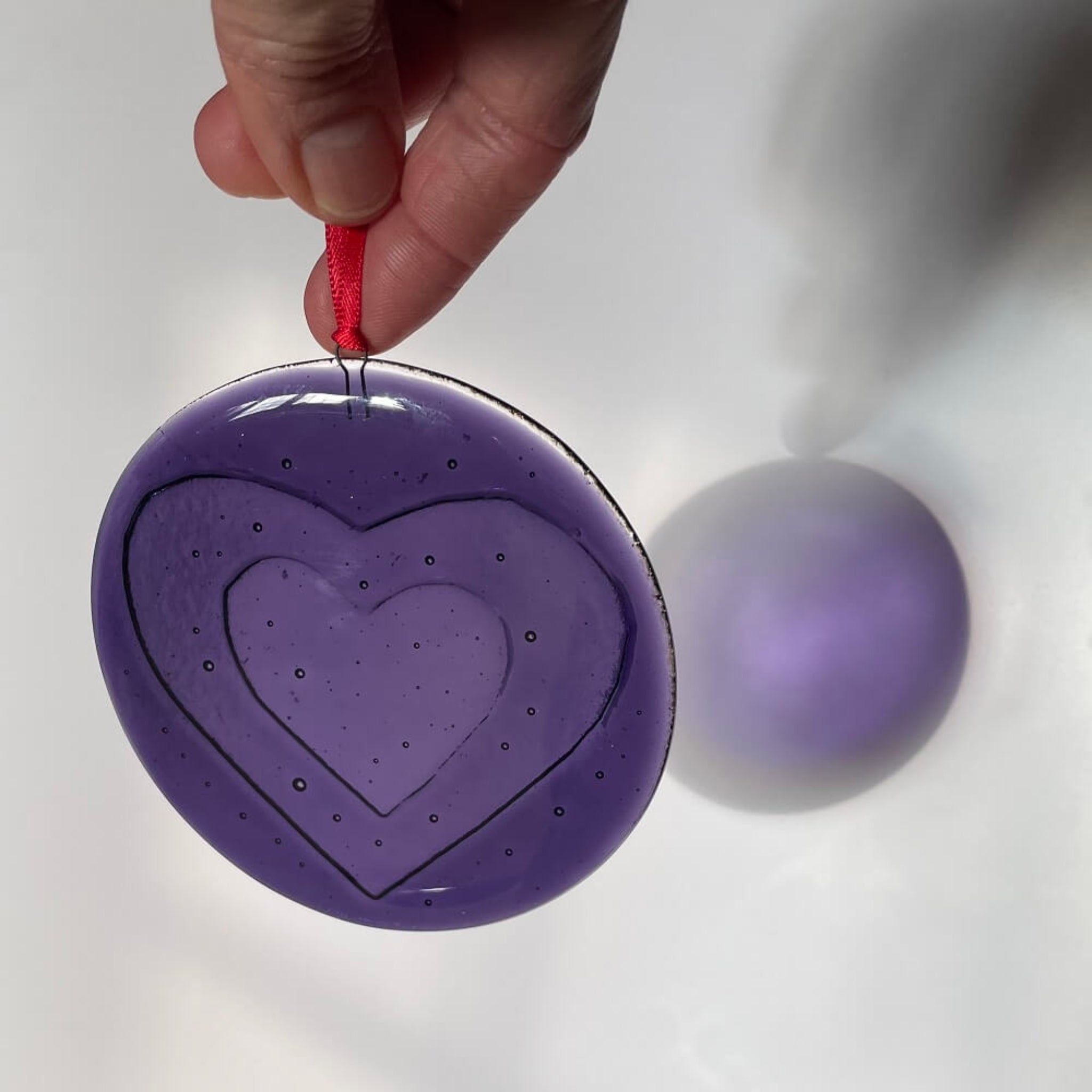 handmade fused glass Purple heart-shaped 'love bomb' hanging decoration / ornament with a red ribbon on a textured white background